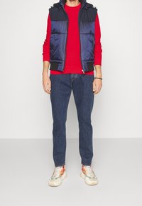 Chaleco acolchado azul marino sobre un suéter rojo, combinado con jeans azules y zapatillas de varios colores. El chaleco tiene un cierre delantero y capucha.