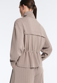 Kvinde med mørkt hår sat op i en hestehale iført beige jakke med nålestriber og matchende bukser, stående med hænderne i lommerne, ryggen vendt mod beskueren.