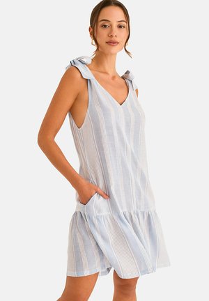 Femme portant une robe sans manches à rayures verticales bleu clair et blanc avec des bretelles nouées aux épaules et des poches, debout devant un fond uni.