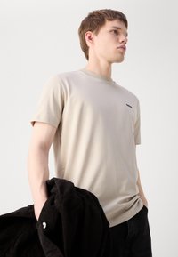 Beige bomulds t-shirt med rund hals og korte ærmer, der har et lille sort logo på venstre bryst. Modellen holder en sort snorejakke.