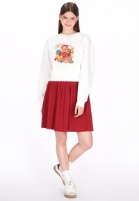Fehér pulóver színes grafikával és szöveggel, piros pliszírozott szoknyával párosítva. A fehér sportcipő teszi teljessé a ruhát.