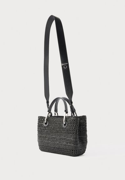 Μαύρη υφαντή τσάντα με δύο κοντά χερούλια και ένα μακρύ ρυθμιζόμενο δερμάτινο λουρί ώμου, παρουσιάζεται μπροστά σε απλό φωτεινό φόντο.