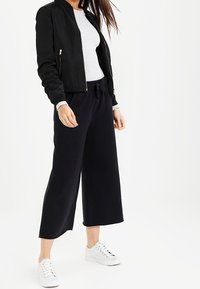 Sort bomberjakke med ribbet manchetter, hvid tætsiddende sweater, løse sorte culottes og hvide sneakers. Glat materiale hele vejen igennem.
