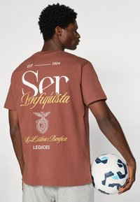 Καφέ T-shirt με κοντό μανίκι, με λευκό και χρυσό κείμενο. Περιέχει ένα γραφικό αετού και λογότυπα ομάδων, κρατούμενο από ένα άτομο με μια μπάλα ποδοσφαίρου.