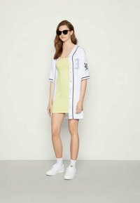 Jersey de beisebol branco com detalhes pretos, usado sobre um vestido amarelo ajustado. A modelo também usa óculos de sol pretos e ténis brancos.