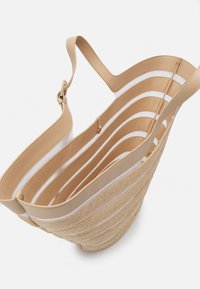 Beige-kudottu kantokassi, jossa on beige nahkaremmi, avoin muotoilu vaakasuorilla raidoilla ja teksturoitu pinta.