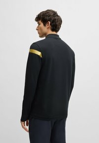 Schwarzes Langarm-Polo mit einem goldenen Streifen auf der Schulter. Besitzt eine lässige Passform und eine weiche Textur. Einfaches, minimalistisches Design.