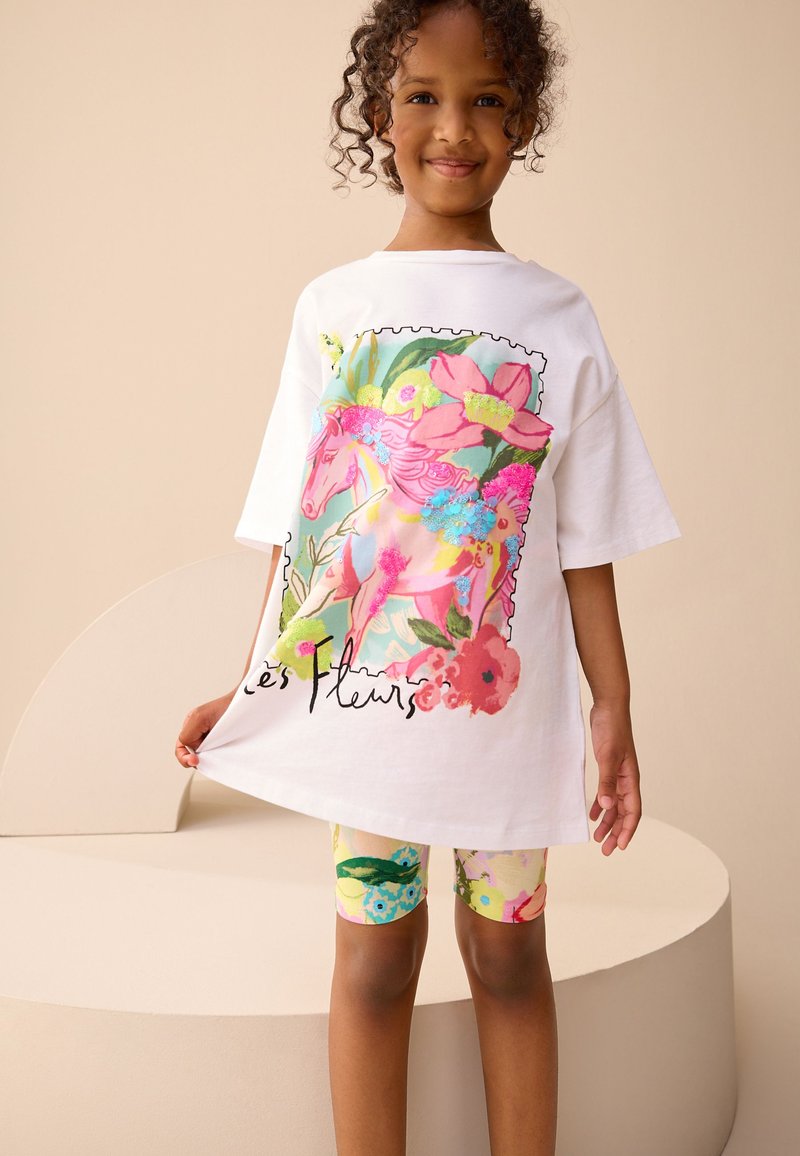 Un t-shirt blanc surdimensionné présente un motif coloré de licorne et de fleurs avec du texte. L'enfant porte des leggings à motifs vifs avec des accents floraux.