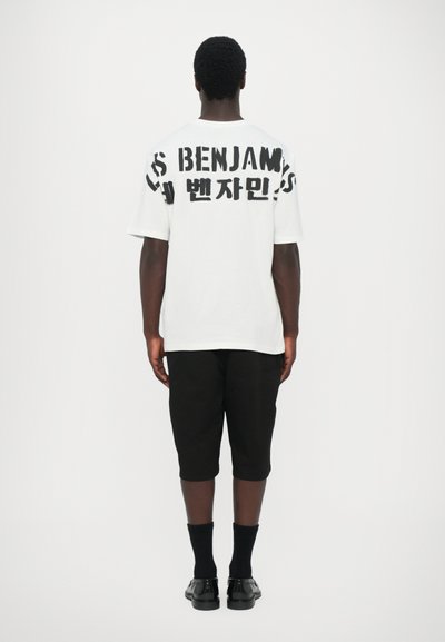 T-shirt en coton blanc avec imprimé graphique noir, associé à un short noir jusqu'aux genoux et des chaussettes noires, porté par une personne de dos.