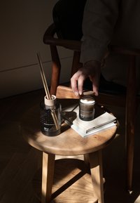Diffuseur en verre avec un couvercle en bois, deux bâtonnets de roseau, et une bougie noire sur un tabouret en bois. Un carnet à couverture douce est posé à côté. La lumière naturelle met en valeur les textures.