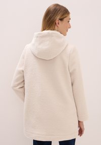 Donna che indossa una giacca in pile beige chiaro e testurizzata con cappuccio, vista di spalle, appoggiata a uno sfondo neutro.