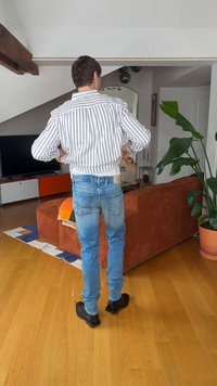 Muž stojí s rukami v bok v bielom pruhovanom saku, modrých džínsoch a čiernych topánkach v obývacej izbe s hrdzavohnedou pohovkou a kvetináčom s rastlinou.