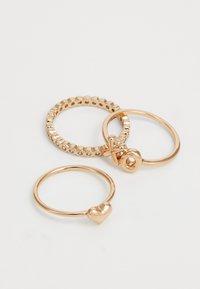 Trois anneaux en or sur une surface blanche : l'un avec un pendentif en forme de cœur, l'autre avec des pierres claires alternées, et le troisième avec des lettres "XO" entrelacées.