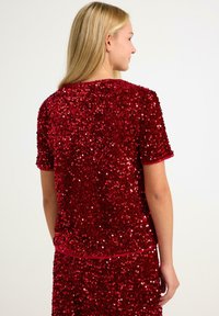 Rød paillet-top med korte ærmer, glat tekstur og rund hals. Har et tætsiddende design, glitrende detaljer og en ensfarvet rød kant.