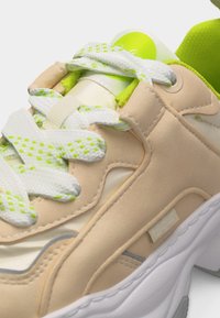 Κοντινό πλάνο μπεζ αθλητικού παπουτσιού με λευκά και πράσινα κορδόνια με σχέδια, πράσινη εσωτερική επένδυση και χοντρή λευκή σόλα σε λευκό φόντο.