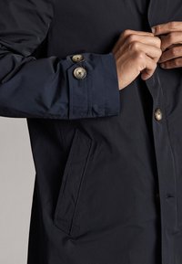 Navy blue, waterproof jacket with a smooth texture, buttoned cuffs, and a side pocket. Features contrasting beige buttons.