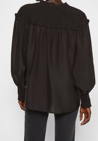 Blouse noire avec un dos smocké, un détail à volants texturé au niveau de l'encolure, et des manches amples, associée à un pantalon foncé.