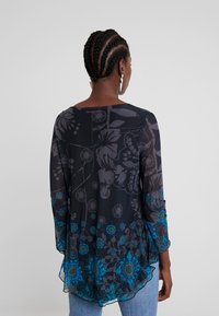 Mujer con el cabello trenzado, vistiendo una blusa negra transparente con estampado floral en azul y turquesa, de pie de espaldas frente a un fondo sencillo.