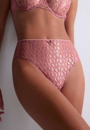 Femme portant des sous-vêtements en dentelle rose transparente taille haute avec motif animalier et soutien-gorge assorti, debout sur un fond neutre.