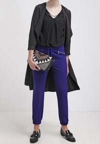 Svart lång jacka över en svart topp, lila byxor med elastiska mudd, och en mönstrad kuvertväska med geometriska mönster. Svarta loafers.