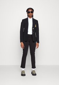 Blazer preto com logótipo dourado, camisa branca de botão, calças pretas justas, óculos de sol oversized pretos e mocassins olivas com solas grossas.