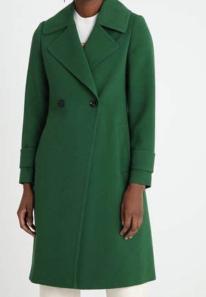 Femme portant un long manteau vert croisé avec de larges revers et des manches retroussées sur un haut blanc et un pantalon clair.