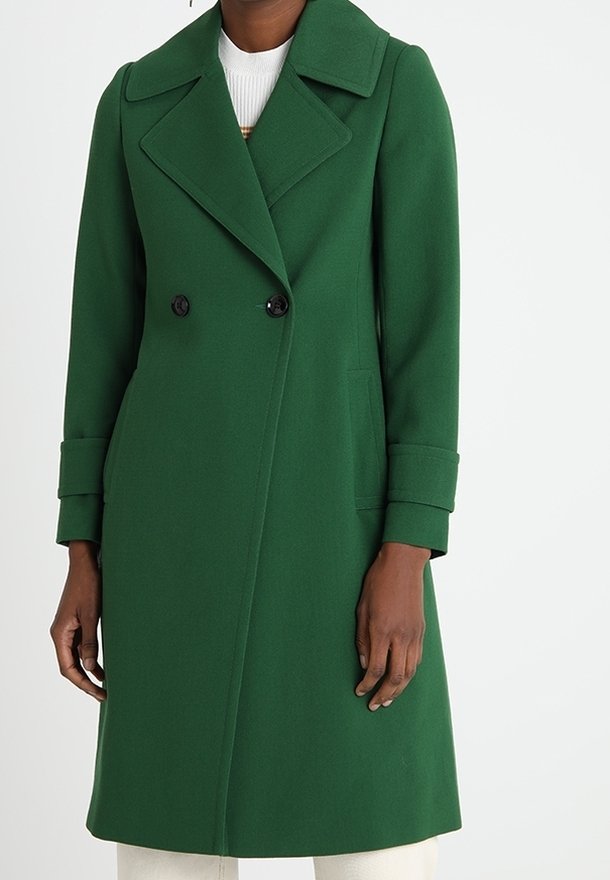 Femme portant un long manteau vert croisé avec de larges revers et des manches retroussées sur un haut blanc et un pantalon clair.