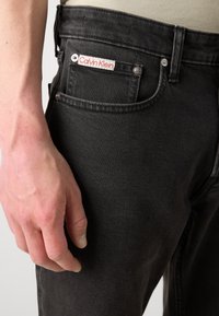 Mano sinistra appoggiata accanto alla tasca di un jeans di denim nero con etichetta bianca Calvin Klein visibile sulla tasca porta monete.