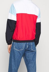 Väripaljetettu takki, jossa on punaisia, valkoisia, vaaleansinisiä ja laivastonsinisiä paneeleja. Takissa on joustava vyötärö ja hihansuut, ja se on suunniteltu sileällä, kiiltävällä pinnalla.