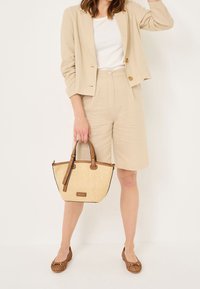 Femme portant un blazer beige et un short, tenant un sac à main tissé beige avec des lanières en cuir marron, et des ballerines tissées marron.