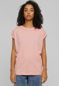 Rosa t-skjorte med korte ermer laget av mykt stoff, med en avslappet passform og rullede ermer, vist sammen med blå denimbukser og en nøytral bakgrunn.