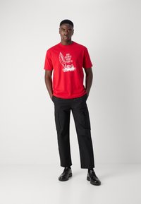 Red cotton t-shirt with a sailboat graphic and white text on the front, paired with black cargo pants and black lace-up shoes.