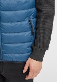 Blå puffväst med horisontella sömmar och sidoficka med blixtlås, tillsammans med en mörkgrå stickad ärm. Texturerat tyg och slät yta.