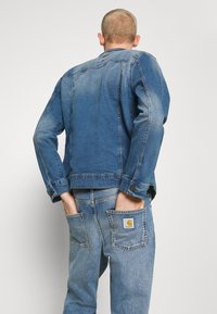 Blå denimjacka med en blekt finish, knappar i ärmsluten och detaljer på bakfickan. Kombinerad med ljusblå jeans med en synlig varumärkeslogotyp.
