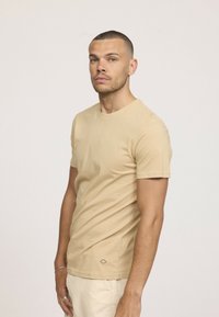 T-shirt beige à manches courtes avec col rond, fabriqué en coton doux. Présente un petit logo près de l'ourlet, coupe ajustée.