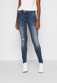 Persona llevando unos pantalones vaqueros ajustados de tiro medio en azul con un pequeño desgarro en la rodilla izquierda y zapatillas blancas, de pie contra un fondo liso.