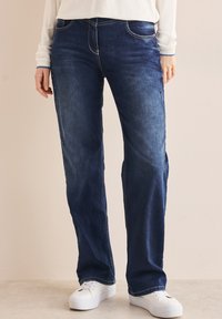 Donkerblauwe denim jeans met een rechte poot, voorzien van voorzakken en een vervaagde afwerking. Gecombineerd met witte sneakers.