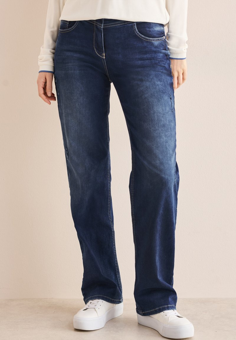 Donkerblauwe denim jeans met een rechte poot, voorzien van voorzakken en een vervaagde afwerking. Gecombineerd met witte sneakers.
