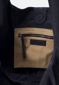 Black suede tote interior featuring a beige fabric zip pocket with a textured surface and a dark label. Secure snap closure visible at the top.