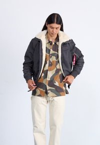 Schwarze Bomberjacke mit Kunstfellkragen, gerippten Bündchen und Seitentaschen. Kombiniert mit einem braun-grünen Camouflage-Hemd und beigen Hosen.