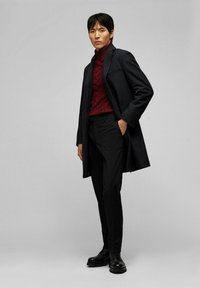 Dunkler Wollmantel mit maßgeschneidertem Schnitt, getragen über einem tiefroten kabelgestrickten Rollkragenpullover. Schwarze Slim-Fit-Hosen und schwarze Schnürstiefel vervollständigen das Outfit.
