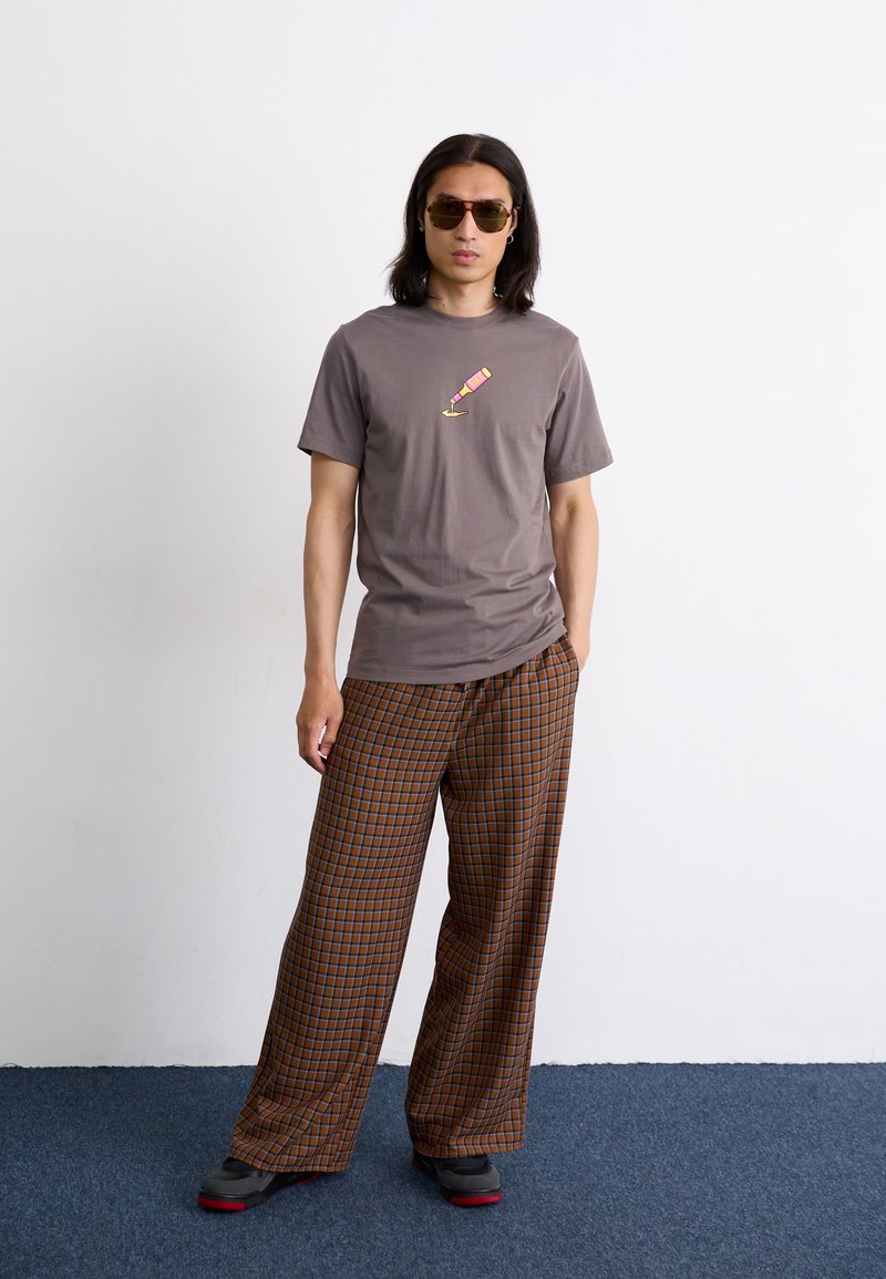 Szary T-shirt z graficznym nadrukiem różowej butelki szampana, w połączeniu z brązowymi spodniami w kratkę o szerokich nogawkach. Noszony z okularami przeciwsłonecznymi i trampkami.