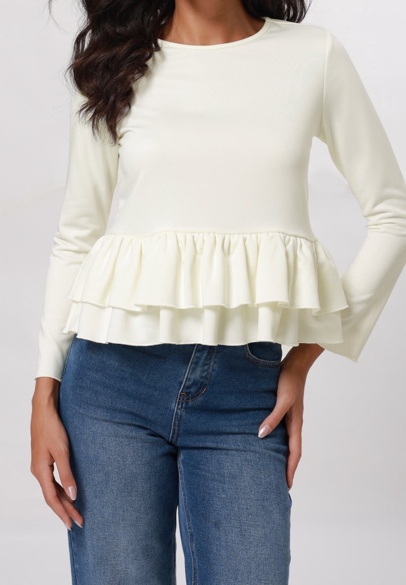 Femme portant un haut crème à manches longues avec un ourlet peplum à volants superposés, associé à un jean en denim bleu, main posée sur la hanche.