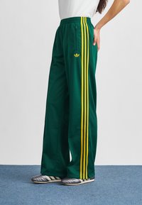 Zielone spodnie sportowe z trzema żółtymi paskami po bokach, wykonane z gładkiej tkaniny, z kieszenią na zamek i logo Adidas.