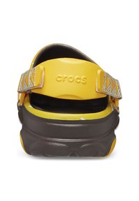 Yellow Crocs with a brown sole, featuring a textured strap with a geometric pattern, open-back design, and cushioned insole for comfort.