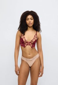 Μπορντό σουτιέν με δαντέλα στα τελειώματα και κυματιστές άκρες με διασταυρούμενες λεπτομέρειες, συνδυασμένο με ροδακινί δαντελένιο στρινγκ με διακοσμητικό φιόγκο.