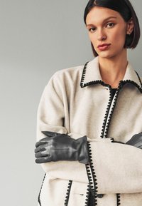 Light beige wool jacket with black scalloped trim, paired with black leather gloves, showcasing smooth texture and structured design.