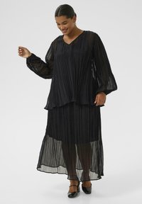 Musta pleatattu mekko kerrostetulla designilla, pitkät hihat ja V-pääntie. Läpinäkyvä kangas ja nilkkapituisuus hienolla tekstuurilla ja liikkeellä.