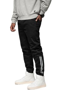 Czarne dresowe spodnie z dwoma białymi paskami wzdłuż nogawek, wykonane z gładkiego materiału. Połączone z białymi butami sportowymi za kostkę i szarym sweatshirtem.