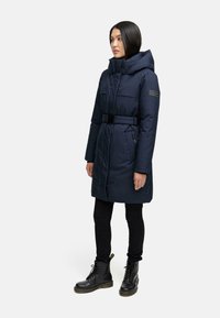 Navy padded coat with a high collar and a belted waist. Features front pockets and a logo patch on the left sleeve. Black skinny pants and boots.